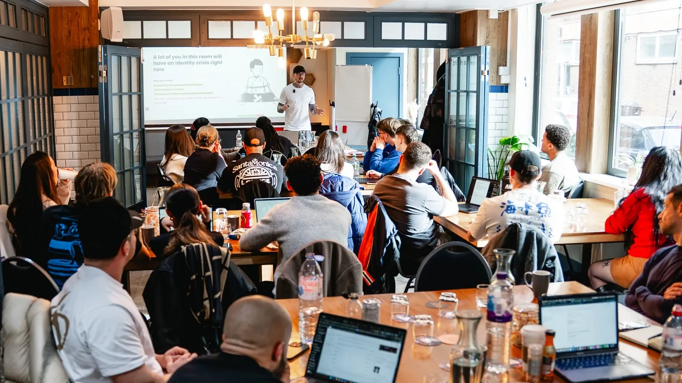 A packed workshop room, clients at tables with laptops out, listening to the session at the front